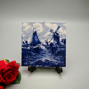 May include: A square ceramic tile with a blue and white Dutch landscape design. The image depicts windmills, a sailing boat, and water under a cloudy sky. The tile is presented on a small black easel.