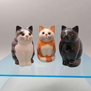 May include: Three ceramic cat figurines standing in a row. The left cat is white with grey and black tabby markings. The centre cat is orange and white with a solid orange face. The right cat is solid black with yellow eyes and a glossy finish.