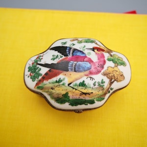 May include: A small, white, ceramic trinket box featuring a vibrant bird design. The bird is depicted with pink, blue, and black plumage, set against green foliage and a small tree. The box is edged with silver-coloured metal trim.