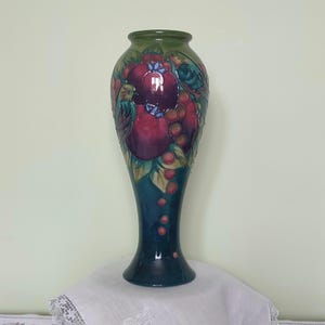 May include: A tall, slender, blue and green ceramic vase with a floral design featuring red and purple fruit and a small bird. The vase has a wide base and a narrow neck.
