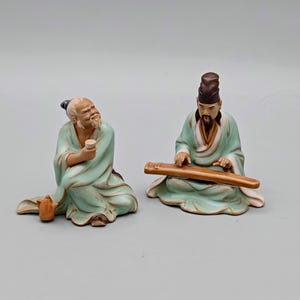 May include: Two ceramic figurines of men in traditional Chinese clothing. One man is holding a cup and a teapot, while the other is playing a stringed instrument.
