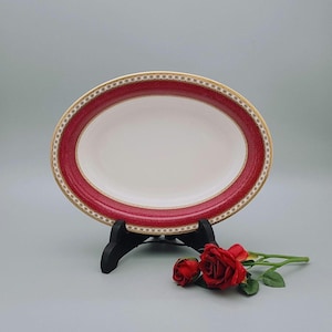 May include: A white oval serving dish with a red and gold rim. The dish is on a black stand and there are two red roses in the foreground.