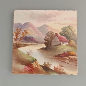 May include: A square ceramic tile featuring a hand-painted landscape scene. The image depicts a small white cottage with a red roof situated beside a calm river, with rolling purple mountains in the background and green grassy banks.