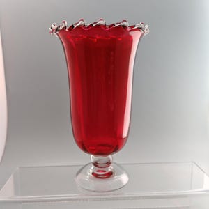 May include: A tall, ruby red glass vase with a flared, ruffled rim featuring a clear glass edge. The vase has a smooth, cylindrical body that tapers slightly towards the base, which is supported by a clear glass pedestal foot and stem.