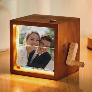 May include: A square wooden music box featuring a light-up front panel that displays a photograph of a smiling couple. The box has a natural brown wood finish and a small wooden hand crank attached to the right side for manual operation.