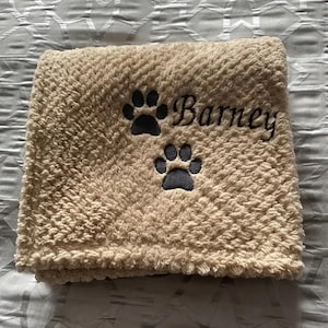 May include: A beige, plush blanket with two dark gray paw prints and the name "Barney" embroidered in black script. The blanket is folded and appears soft and cozy, perfect for pets.
