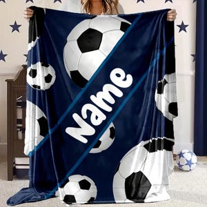 May include: A navy blue blanket with a soccer ball pattern. The blanket has a diagonal stripe with the white word "Name" printed on it. Several soccer balls are scattered across the blanket.