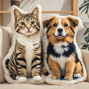May include: Two decorative pillows shaped like a cat and a dog. The cat pillow has a tabby pattern with brown and black stripes. The dog pillow features a brown, black, and white color scheme, with a smiling expression. Both pillows are on a beige sofa.