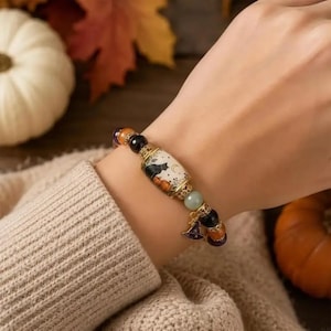 Peut inclure: Un bracelet de perles avec une perle cylindrique centrale ornée d'une illustration de chat noir et de citrouille orange, agrémenté de perles rondes noires, orange et vert pâle, et d'une petite breloque en forme de chapeau de sorcière.