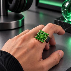 May include: A gold-toned ring featuring a prominent green circular emblem with a central spiral design, surrounded by a rectangular frame filled with small, sparkling green stones. The band of the ring is a solid, dark matte black color.
