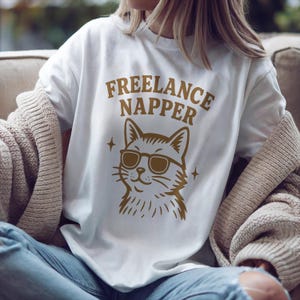 Cartoon-style cat wearing oversized sunglasses with a content smile. Above the cat, bold text reads “Freelance Napper.” The background is transparent, and the illustration includes sparkles around the character for added charm. Provided as a high-resolution PNG file, perfect for sublimation or crafting. No physical product included—digital download only.