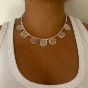 Peut inclure: Un collier délicat composé de petites perles roses et de pendentifs circulaires plats et irisés. Le collier est porté sur un débardeur blanc, créant un contraste de textures et de couleurs. Le collier est un bijou.