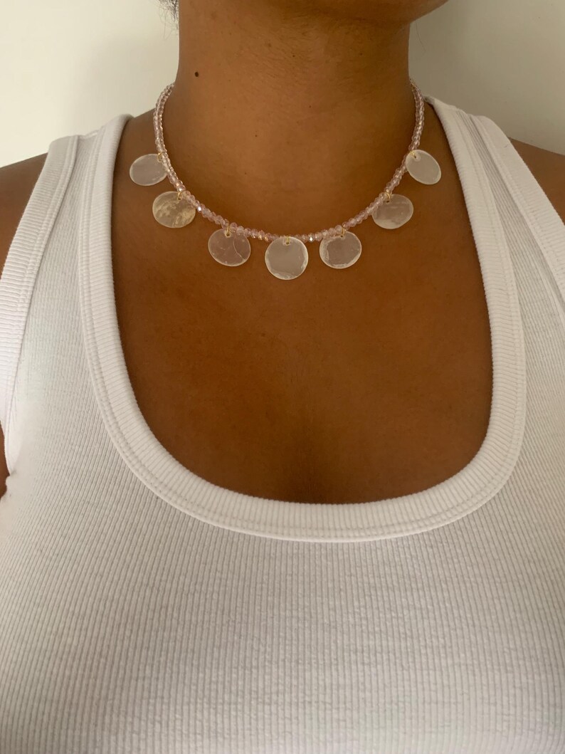 Pode incluir: Um colar delicado com uma corrente de pequenas contas rosa e pingentes circulares planos e iridescentes. O colar &eacute; usado sobre uma regata branca, criando um contraste de texturas e cores. O colar &eacute; uma joia.