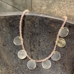 Pode incluir: Um colar delicado com um fio de pequenas contas rosa claro. O colar &eacute; adornado com v&aacute;rios pingentes planos, redondos e iridescentes. Os pingentes s&atilde;o presos ao fio de contas com pequenos conectores dourados. O colar &eacute; exibido em uma superf&iacute;cie escura e texturizada.