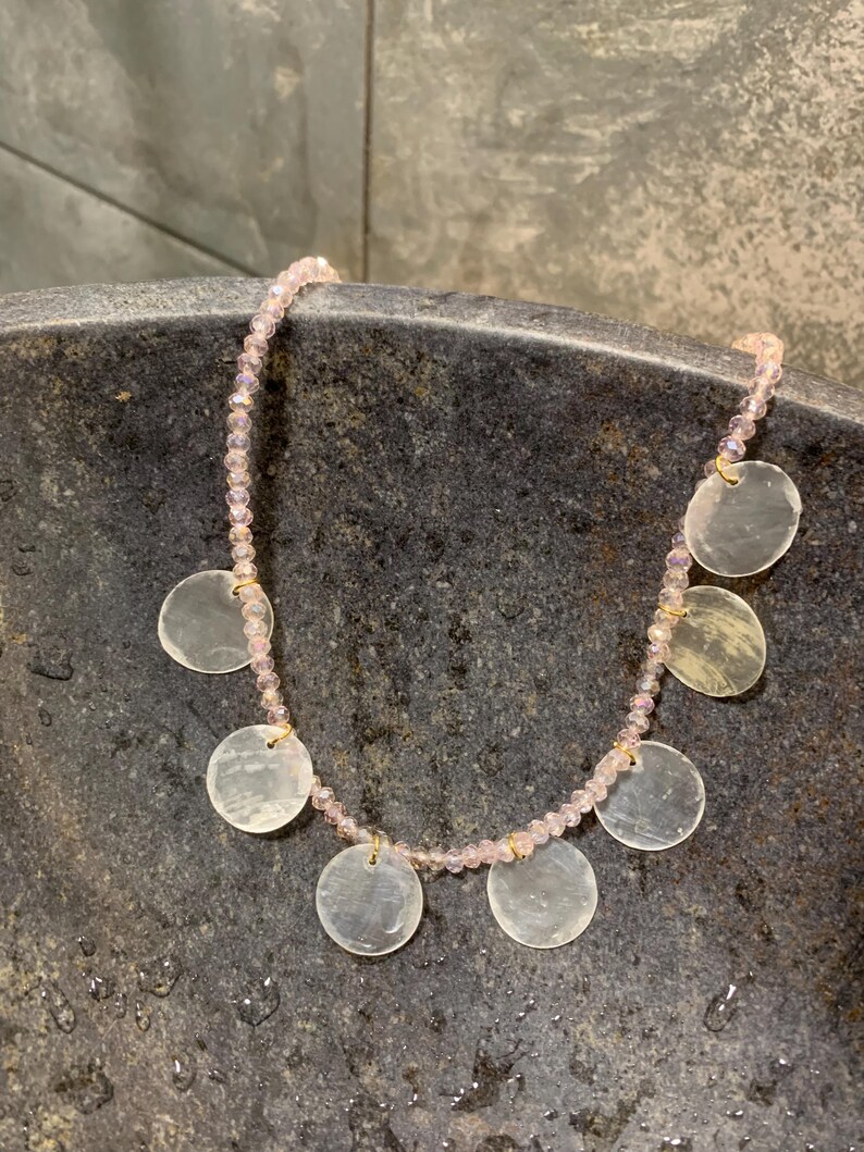 Pode incluir: Um colar delicado com um fio de pequenas contas rosa claro. O colar &eacute; adornado com v&aacute;rios pingentes planos, redondos e iridescentes. Os pingentes s&atilde;o presos ao fio de contas com pequenos conectores dourados. O colar &eacute; exibido em uma superf&iacute;cie escura e texturizada.