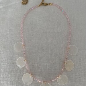 Pode incluir: Um colar delicado com uma corrente de pequenas contas rosa claro. O colar &eacute; adornado com seis pingentes redondos de concha branca e uma corrente dourada com um pingente de sol.