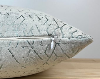 May include: A light gray throw pillow with a geometric pattern in shades of green. The pillow has a zipper closure.