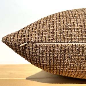 Puede incluir: Primer plano de un cojín texturizado marrón y beige. El cojín tiene un aspecto tejido con cierre de cremallera. El cojín es de color neutro y complementaría varios estilos de decoración del hogar.