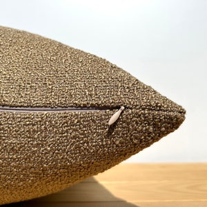 May include: Close-up of a brown throw pillow with a zipper closure. The pillow is made of a textured fabric.