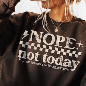 K&ouml;nnte beinhalten: Ein dunkelbraunes Sweatshirt mit dem wei&szlig;en Text "NOPE not today", einem Schachbrettmuster und einem Blitz-Grafik. Das Sweatshirt hat auch den Text "and tomorrow's not looking good either". Eine schwarze Tasse wird gehalten.