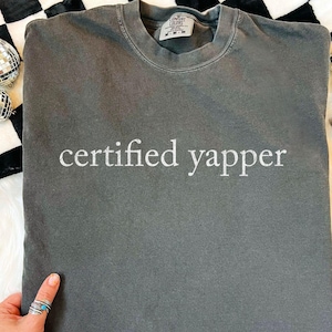 Op de afbeelding: Een grijs Comfort Colors crewneck sweatshirt met de witte tekst "certified yapper". Het shirt is opgevouwen en gepresenteerd op een zwart-wit geblokt oppervlak. Decoratieve discoballen en sieraden zijn zichtbaar.