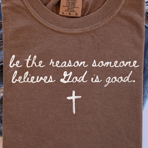 Op de afbeelding: Een bruin t-shirt met de tekst "be the reason someone believes God is good" en een kruis. Het shirt is opgevouwen naast een spijkerbroek.