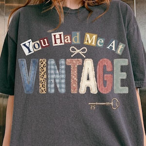 以下が含まれることがあります： 「You Had Me At Vintage」の文字がコラージュ風にデザインされたダークグレーのTシャツ。「Vintage」の文字は様々な模様と色で表現されています。「Vintage」の下には小さな鍵のグラフィックがあります。