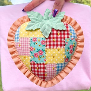 Può includere: Una borsa tote rosa chiaro con un grande design a fragola applicato. La fragola è realizzata in tessuto patchwork con vari motivi, tra cui quadretti e stampe floreali, con un bordo a balze e dettagli di foglie verdi. La borsa è tenuta su uno sfondo verde.