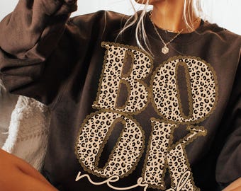 Könnte beinhalten: Ein braunes Sweatshirt mit dem Text "BOOK nerd" in einem Leopardenmuster, umrandet von einer dekorativen Bordüre. Eine schwarze Tasse wird in der Nähe des Gesichts gehalten.