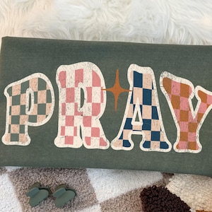 May include: A green t-shirt with the word "PRAY" in a retro font. The letters have a checkered pattern in pink, blue, and brown. A brown and white cowboy boot is visible in the corner.