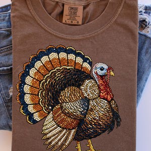 May include: A brown Comfort Colors t-shirt featuring a detailed turkey design with a vibrant tail in shades of blue, orange, and brown. The turkey has a red wattle and a blue head. The shirt is paired with blue denim jeans.