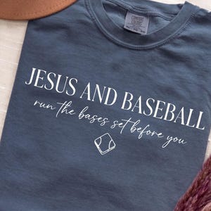 May include: A slate blue t-shirt with the words "JESUS AND BASEBALL" in white, along with the phrase "run the bases set before you" and a baseball graphic. A brown hat is partially visible in the upper left corner.