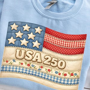 Könnte beinhalten: Hellblaues Sweatshirt mit einem patriotischen Design. Das Design zeigt eine stilisierte amerikanische Flagge mit Sternen, Streifen und dem Text "USA 250". Das Flaggen-Design ist in Rot, Weiß und Blau gehalten.
