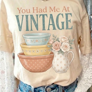 May include: Beige t-shirt with the text "You Had Me At Vintage" in pink and teal. The shirt features a graphic of three decorative bowls and a floral pitcher with roses. The shirt has short sleeves and is worn over a white lace long sleeve shirt.