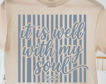 Könnte beinhalten: Cremefarbenes T-Shirt mit blauem Streifendesign und dem Schriftzug "it is well with my soul" in Schreibschrift. Das Shirt hängt an einem Holzkleiderbügel und zeigt seinen lässigen Stil und seine inspirierende Botschaft.
