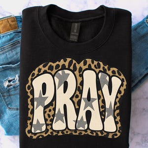 May include: Black crew neck sweatshirt featuring the word "PRAY" in a retro font, filled with grey stars. The word is outlined with a leopard print design. The sweatshirt is displayed with a pair of blue jeans.