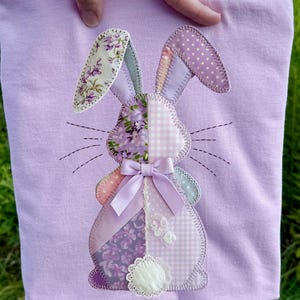 Op de afbeelding: Een lichtpaars T-shirt met een patchwork konijnontwerp. Het konijn heeft bloemen-, stippen- en ruitpatronen in paars, roze en wit. Een lavendelkleurig lint en een witte kanten staart maken het ontwerp compleet.
