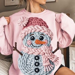 May include: A light pink sweatshirt featuring a knitted snowman design. The snowman has a blue body, a pink hat and scarf, an orange nose, and black button eyes and buttons. Brown twig arms complete the design.