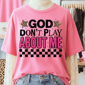 May include: A pink t-shirt with the text "GOD DON'T PLAY ABOUT ME" in black and pink. The design includes leopard print stars and a black and white checkered pattern at the bottom.