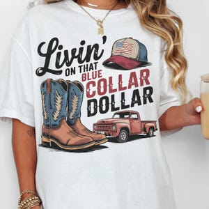 May include: White t-shirt with a graphic design featuring a pair of cowboy boots, a pickup truck, and a baseball cap with an American flag. The text on the shirt reads "Livin' on that Blue Collar Dollar."