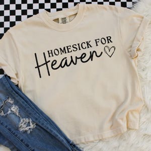 May include: Cream-colored t-shirt with the text "Homesick for Heaven" in black script and a heart symbol. The shirt is paired with blue jeans with distressed details. The shirt has a relaxed fit and rolled sleeves.