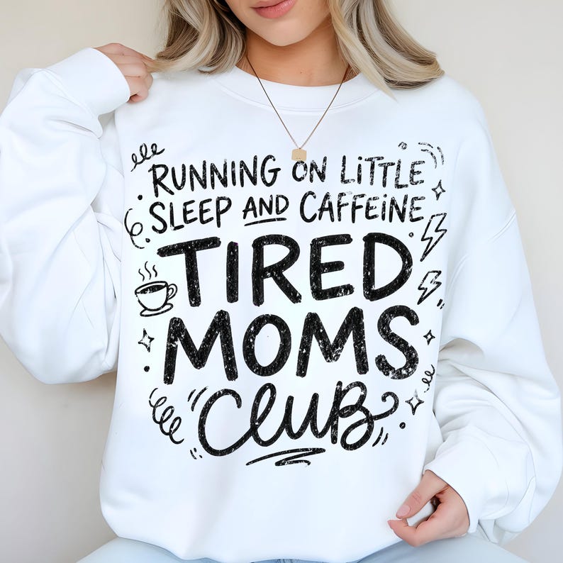 May include: White sweatshirt with the text "Running on Little Sleep and Caffeine, Tired Moms Club" in black. The design includes a coffee cup, stars, and lightning bolts. A casual, comfortable top for moms.