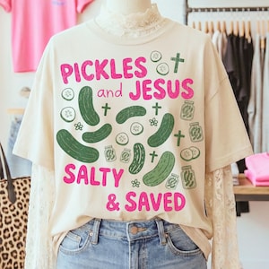 Könnte beinhalten: Cremefarbenes T-Shirt mit den pinkfarbenen Worten "PICKLES and JESUS SALTY & SAVED". Das Shirt zeigt grüne Gurken-Illustrationen und Kreuze. Das Shirt wird auf einer Schaufensterpuppe präsentiert.