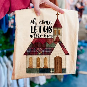 Puede incluir: Un artículo de tela beige con un diseño de iglesia. La iglesia tiene un tejado rojo, una puerta marrón y una cruz. El texto "oh come LET US adore him" está impreso en negro. El artículo está sostenido por una persona.