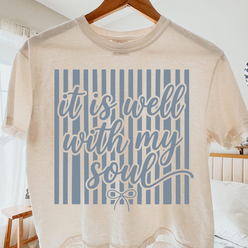 May include: Cream-colored t-shirt with a blue striped design and the text "it is well with my soul" in a cursive font. The shirt is hanging on a wooden hanger, showcasing its casual style and inspirational message.