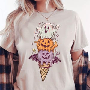 Puede incluir: Camiseta beige con un diseño de cono de helado de Halloween. El diseño incluye un fantasma, una calabaza de Halloween y una bola de helado morada con alas de murciélago, todo sobre un cono de gofre. El gráfico es en tonos naranja, morado y blanco.