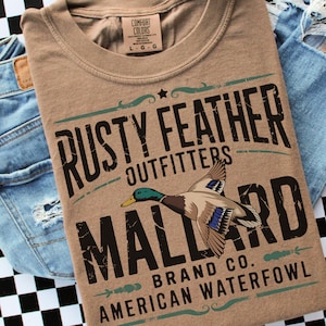 May include: A tan Comfort Colors t-shirt with a mallard duck graphic and the text "RUSTY FEATHER OUTFITTERS MALLARD BRAND CO. AMERICAN WATERFOWL." The shirt is displayed with blue denim jeans.