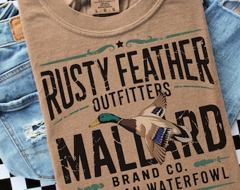 May include: A tan Comfort Colors t-shirt featuring a graphic of a mallard duck and the text "RUSTY FEATHER OUTFITTERS MALLARD BRAND CO. AMERICAN WATERFOWL." The shirt is paired with blue denim jeans.