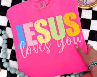 May include: A bright pink t-shirt with the words "JESUS loves you" printed in colorful block letters. The shirt is a crew neck style and appears to be made of a soft material. The text is in blue, pink, yellow, and green.