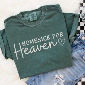 May include: A green t-shirt with the words "Homesick For Heaven" in white script and a heart symbol. The shirt is folded and placed next to a pair of blue jeans.
